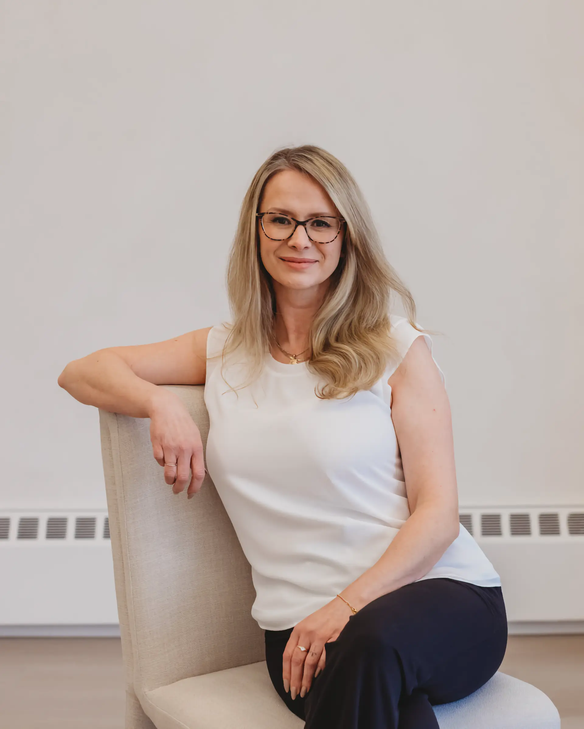 Sarah Hope, a clinical counsellor wearing glasses, smiling warmly with her arm resting on a cream linen chair