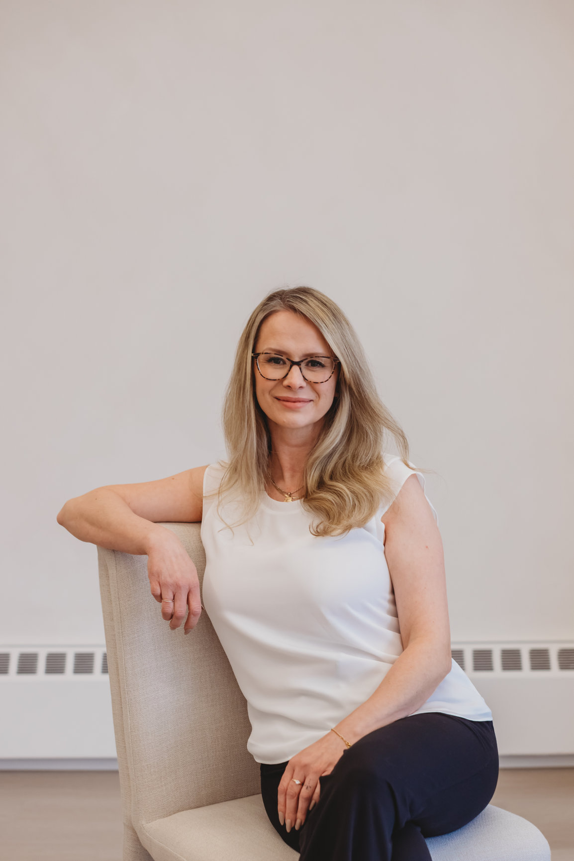 Sarah Hope, clinical counsellor, seated and smiling warmly in glasses, resting on a chair armrest in a bright room